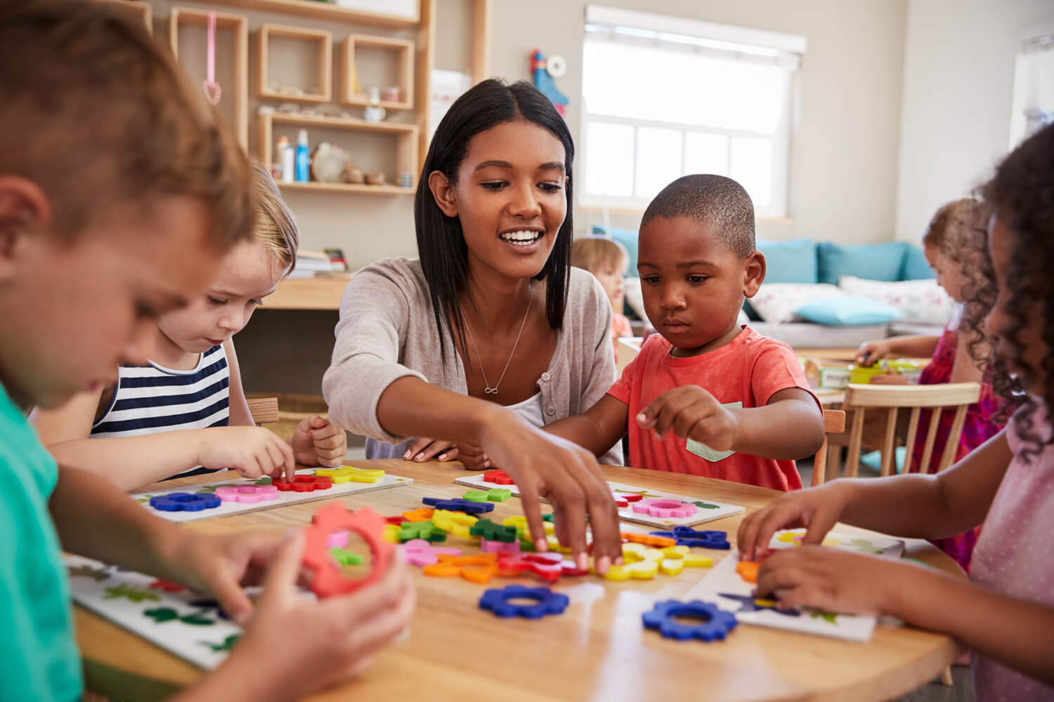Preschool teacher assisting multiple children around a table with shapes